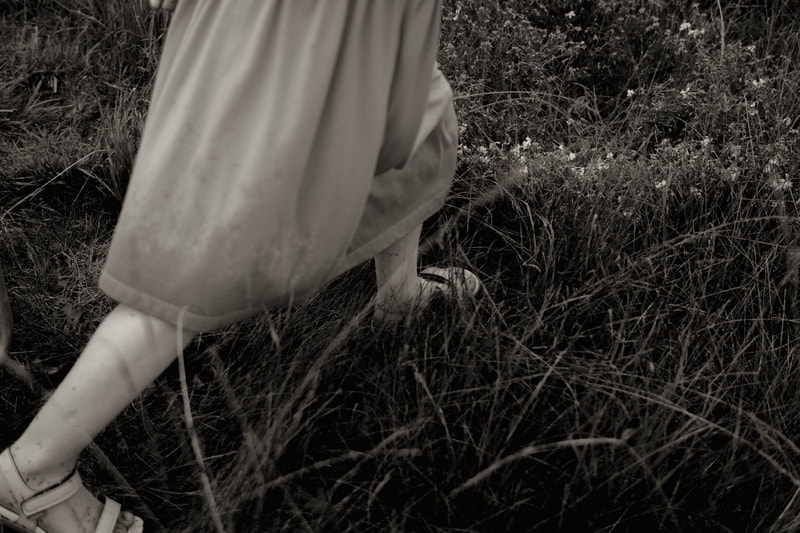 a black and white photograph of a little girls feet and legs as she walks through a meadow, by Minnesota-based photographer Kayla Schiltgen